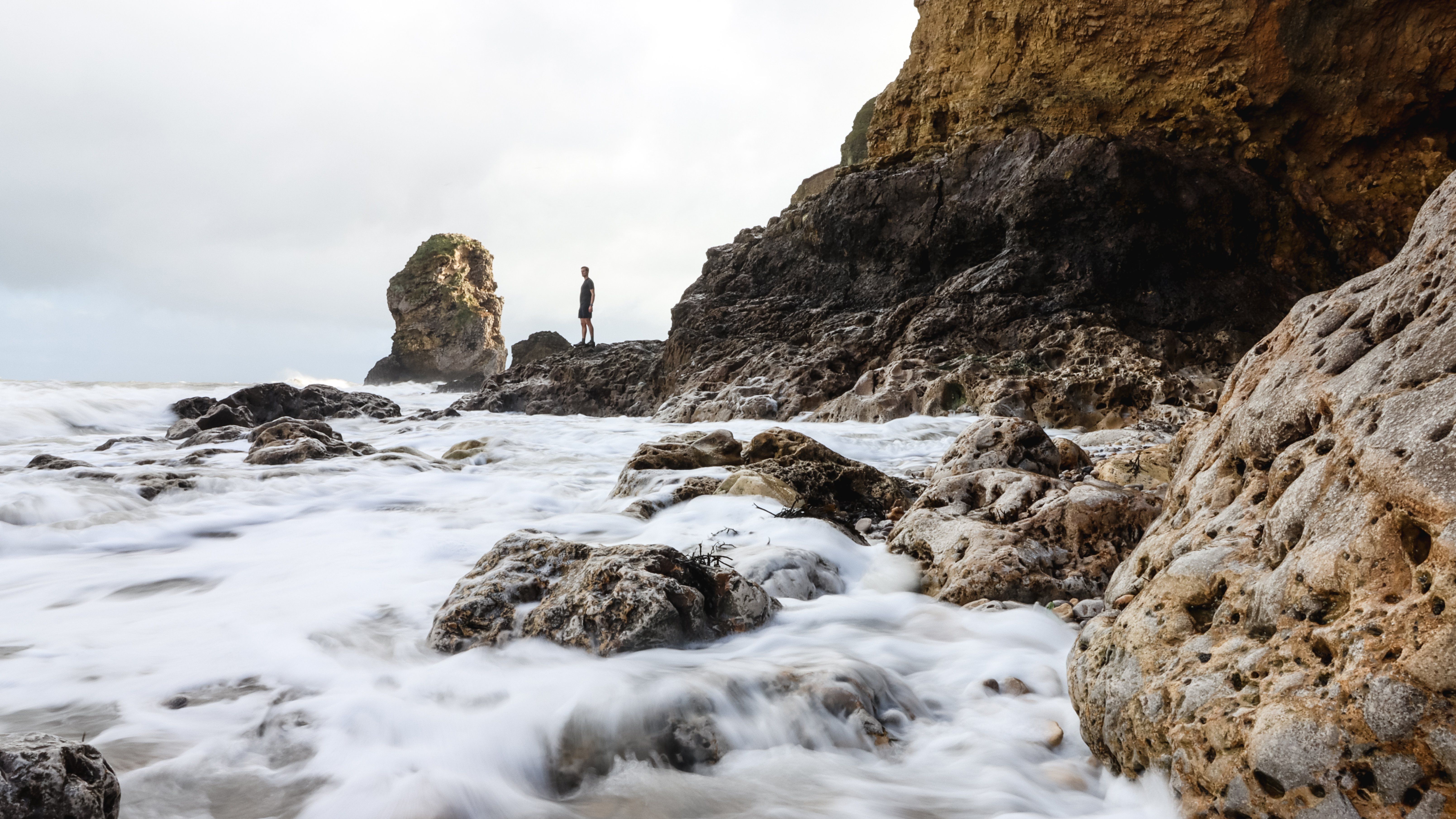 A man stands on rocks by the coast, with the moving waves blurred and softened by a long exposure.